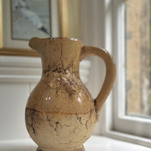 May include: A beige ceramic jug with a textured surface and a handle. The jug has a rustic, vintage look.
