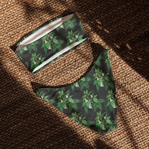 May include: A two-piece bikini set featuring a bandeau top and high-waisted bottoms. The fabric is dark gray and decorated with a repeating botanical pattern of green leaves and small white trumpet-shaped flowers throughout the entire garment.
