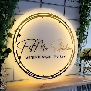 May include: A circular, backlit wall sign featuring the text FitMe Studio and Sağıklı Yaşam Merkezi in black lettering. The sign has a gold-toned circular frame with decorative accents and emits a warm, soft glow from behind the acrylic surface.