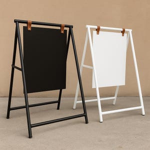 May include: Two metal A-frame signs, one black and one white, with rectangular panels. The signs have a minimalist design with brown leather straps at the top. The black sign is on the left, and the white sign is on the right.