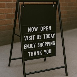 May include: Black metal A-frame sign with the words "NOW OPEN VISIT US TODAY ENJOY SHOPPING THANK YOU" in white block letters. The sign is displayed on a gray concrete surface in front of a brick wall.