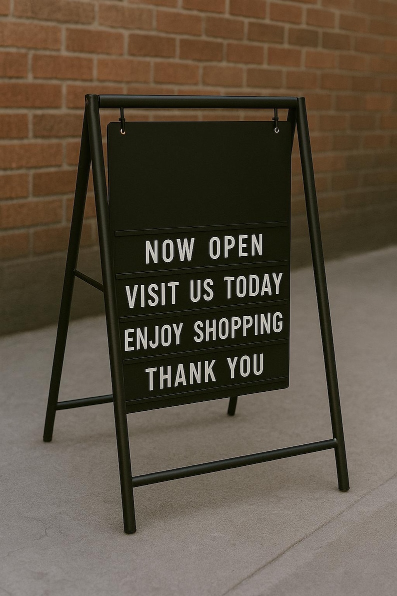 May include: Black metal A-frame sign with the words "NOW OPEN VISIT US TODAY ENJOY SHOPPING THANK YOU" in white block letters. The sign is displayed on a gray concrete surface in front of a brick wall.