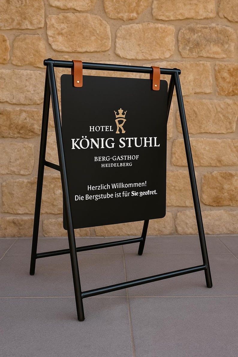May include: Black metal A-frame sign with the text "HOTEL K&Ouml;NIG STUHL" in gold lettering. The sign also includes the text "BERG-GASTHOF HEIDELBERG" and a welcome message. The sign is held by brown leather straps.