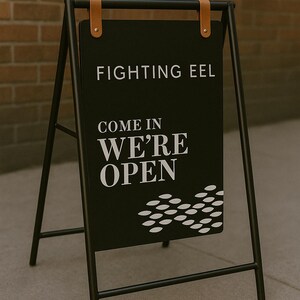 May include: Black sandwich board sign with the text "FIGHTING EEL COME IN WE'RE OPEN" in white. The sign is held by a black metal frame with brown leather straps at the top. A brick wall is in the background.