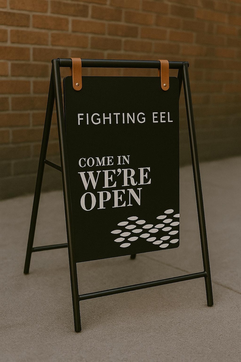 May include: Black sandwich board sign with the text "FIGHTING EEL COME IN WE'RE OPEN" in white. The sign is held by a black metal frame with brown leather straps at the top. A brick wall is in the background.