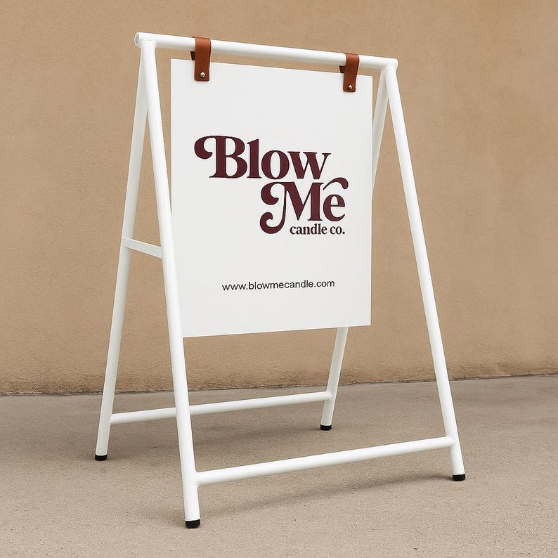 May include: White metal A-frame sign with a white rectangular sign. The sign has the text "Blow Me candle co." in burgundy script. The sign is held in place by brown leather straps. The website address is printed at the bottom.