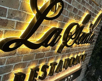 Może przedstawiać: Ceglana ściana z szyldem "La Scala Restaurant" w czarnych, podświetlanych literach. Znak zawiera również "EST. 1977" w białych literach. Litery są obrysowane ciepłą, żółtą poświatą.