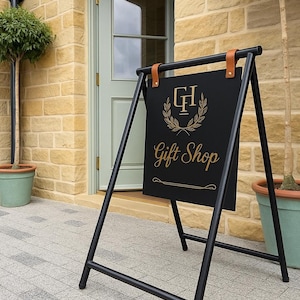 May include: Black metal A-frame sign with the words "Gift Shop" in gold script. The sign has a gold emblem and is attached with brown leather straps. The sign is in front of a building with a light green door.