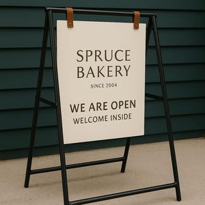 May include: A-frame sign with a black metal frame and a cream-colored sign. The sign reads "SPRUCE BAKERY SINCE 2004 WE ARE OPEN WELCOME INSIDE". The sign is held in place with brown leather straps.