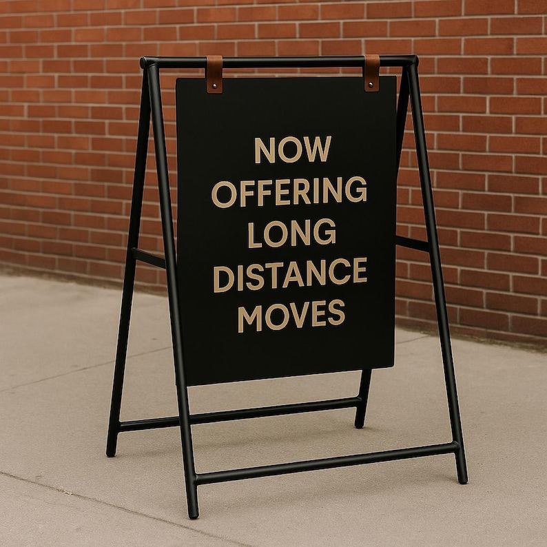 May include: A-frame sign with a black chalkboard and black metal frame. The sign reads "NOW OFFERING LONG DISTANCE MOVES" in a sans-serif font. The sign is attached to the frame with brown leather straps.
