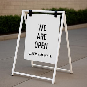 May include: A white A-frame sign with black text that reads "WE ARE OPEN. COME IN AND SAY HI." The sign is supported by a white metal frame and black hinges. The sign is placed on a gray concrete surface.