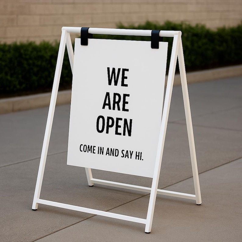 May include: A white A-frame sign with black text that reads "WE ARE OPEN. COME IN AND SAY HI." The sign is supported by a white metal frame and black hinges. The sign is placed on a gray concrete surface.