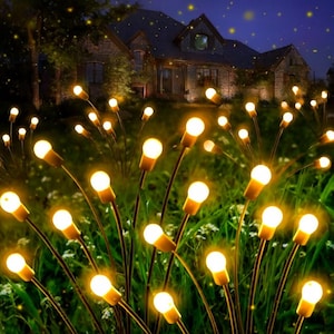 May include: Outdoor decorative lights with warm white bulbs. The lights are arranged in a garden setting, with a house in the background. The bulbs are on slender stems, creating a firefly-like effect. The scene is illuminated at night.