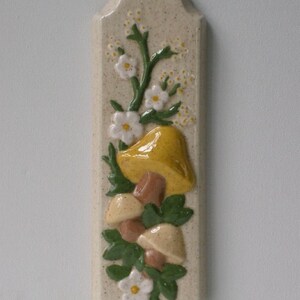 May include: A rectangular ceramic wall plaque with a beige speckled background. The plaque features a raised relief design of a yellow mushroom, brown mushrooms, white flowers, and green leaves. The design is centered on the plaque.