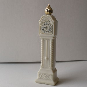 May include: Ivory-colored miniature grandfather clock with a gold-colored top and clock face. The clock face has Roman numerals and gold trim. The clock has a decorative rope-like design on the sides and a detailed base. The clock is a decorative item.