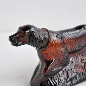 May include: A brown glass figurine of a dog, possibly a setter or spaniel, in a resting pose. The figurine has detailed fur and facial features, with a collar around its neck. The dog is resting on a base with floral details.