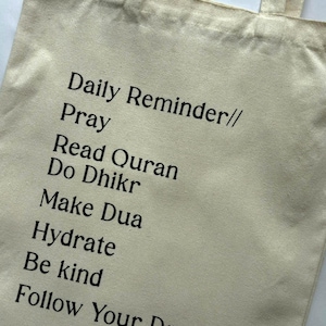 May include: Beige tote bag with black text that reads "Daily Reminder// Pray Read Quran Do Dhikr Make Dua Hydrate Be kind Follow Your Dreams". The bag has two handles.