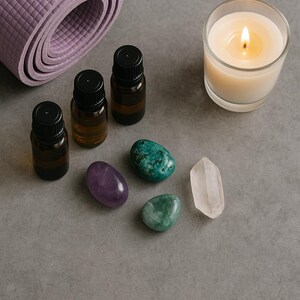 May include: A rolled-up lavender yoga mat, essential oil bottles, a lit candle in a glass, and polished gemstones in shades of purple, green, and clear. The items are arranged on a gray surface, suggesting a wellness or meditation theme.