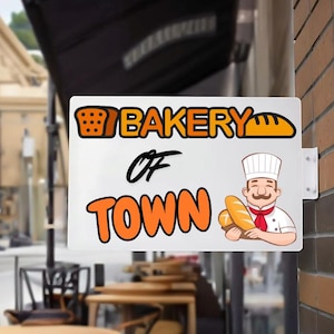 May include: A white rectangular sign for a bakery, mounted on a brick wall. The sign features the words "Bakery of Town" in orange and black, along with illustrations of bread and a chef holding loaves.