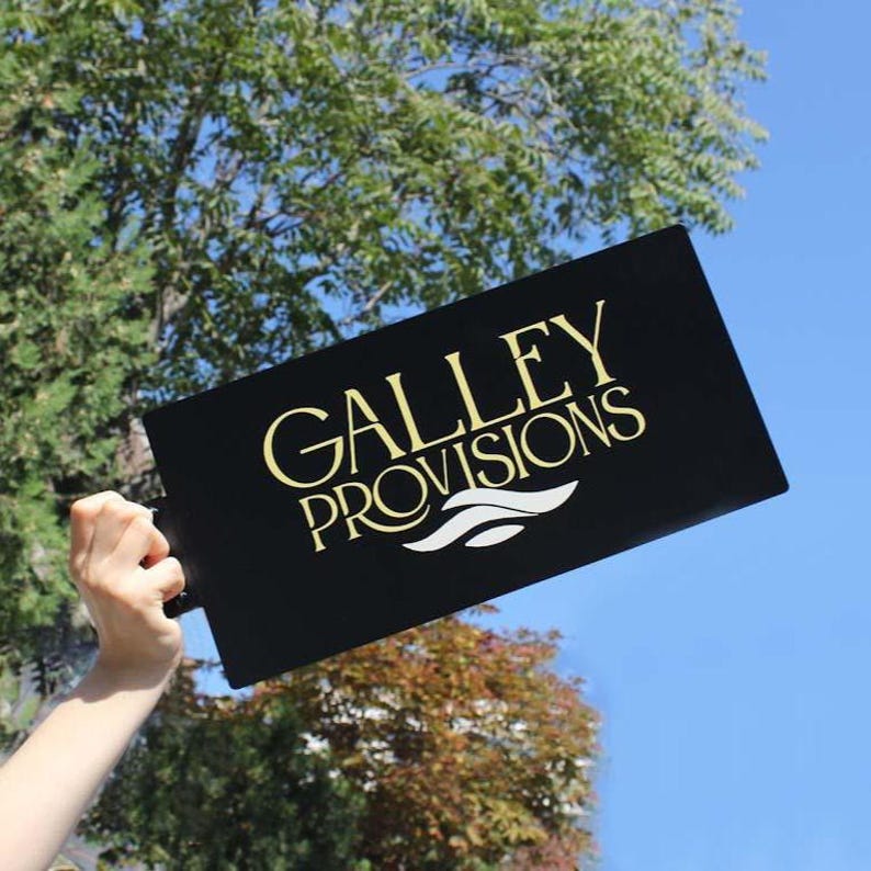 Peut inclure: Un panneau rectangulaire noir avec les mots "GALLEY PROVISIONS" en lettres dor&eacute;es et un motif de vagues blanches. Le panneau est tenu devant un ciel bleu et un feuillage vert.