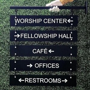 Peut inclure: Panneau directionnel noir avec texte blanc indiquant les emplacements : Worship Center, Fellowship Hall, Caf&eacute;, Offices et Restrooms. Le panneau est suspendu &agrave; une barre m&eacute;tallique noire. Le fond est vert.