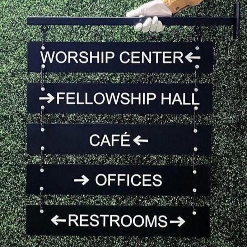 Peut inclure: Panneau directionnel noir avec texte blanc indiquant les emplacements : Worship Center, Fellowship Hall, Caf&eacute;, Offices et Restrooms. Le panneau est suspendu &agrave; une barre m&eacute;tallique noire. Le fond est vert.