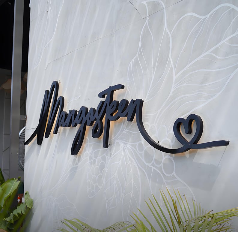 K&ouml;nnte beinhalten: Schwarzes Metallschild mit der Aufschrift "MangosTeen" in Schreibschrift, verziert mit einem herzf&ouml;rmigen Schn&ouml;rkel. Das Schild ist an einer hellgrauen Wand mit wei&szlig;em Blumenmuster angebracht. Gr&uuml;ne Pflanzen sind im Vordergrund zu sehen.