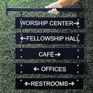 Peut inclure: Panneau directionnel noir avec texte blanc indiquant les emplacements : Worship Center, Fellowship Hall, Caf&eacute;, Offices et Restrooms. Le panneau est suspendu &agrave; une barre horizontale noire et tenu par une main gant&eacute;e sur fond vert.