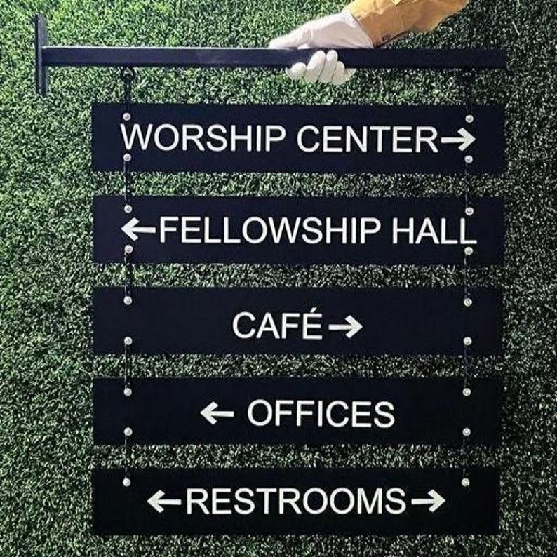 Peut inclure: Panneau directionnel noir avec texte blanc indiquant les emplacements : Worship Center, Fellowship Hall, Caf&eacute;, Offices et Restrooms. Le panneau est suspendu &agrave; une barre horizontale noire et tenu par une main gant&eacute;e sur fond vert.