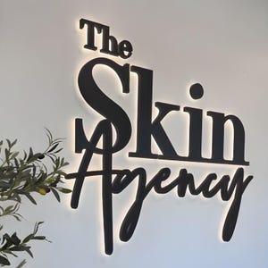 K&ouml;nnte beinhalten: Schwarz beleuchtetes Schild mit der Aufschrift "The Skin Agency" in einer modernen, eleganten Schriftart. Das Schild ist an einer wei&szlig;en Wand angebracht, mit einem subtilen Gl&uuml;hen, das von hinter den Buchstaben ausgeht. Ein Zweig mit Gr&uuml;n ist links zu sehen.