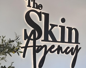 May include: Black illuminated sign that reads "The Skin Agency" in a modern, elegant font. The sign is mounted on a white wall, with a subtle glow emanating from behind the letters. A sprig of greenery is visible on the left.