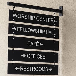 Peut inclure: Panneau directionnel noir avec texte blanc indiquant les emplacements : Worship Center, Fellowship Hall, Caf&eacute;, Offices et Restrooms. Le panneau est suspendu &agrave; une barre m&eacute;tallique noire et mont&eacute; sur un mur textur&eacute;.