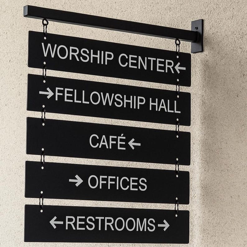 Peut inclure: Panneau directionnel noir avec texte blanc indiquant les emplacements : Worship Center, Fellowship Hall, Caf&eacute;, Offices et Restrooms. Le panneau est suspendu &agrave; une barre m&eacute;tallique noire et mont&eacute; sur un mur textur&eacute;.