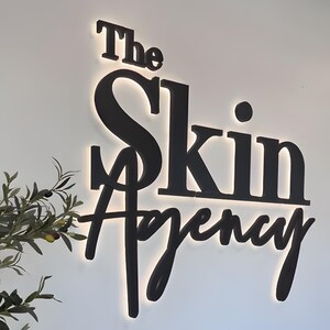 K&ouml;nnte beinhalten: Ein schwarzes Schild mit der Aufschrift &bdquo;The Skin Agency&ldquo; in einer stilisierten Schriftart. Das Schild ist von hinten beleuchtet und erzeugt einen Leuchteffekt. Ein Zweig mit Gr&uuml;n ist unten links zu sehen.