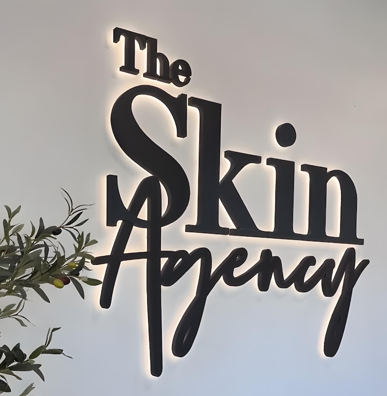 K&ouml;nnte beinhalten: Ein schwarzes Schild mit der Aufschrift &bdquo;The Skin Agency&ldquo; in einer stilisierten Schriftart. Das Schild ist von hinten beleuchtet und erzeugt einen Leuchteffekt. Ein Zweig mit Gr&uuml;n ist unten links zu sehen.