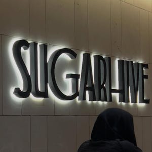 K&ouml;nnte beinhalten: Das Wort "SUGARHIVE" in gro&szlig;en, schwarzen, beleuchteten Buchstaben vor einer neutralen Wand. Die Buchstaben haben einen 3D-Effekt mit hellem Licht umrandet. Eine Person in einer schwarzen Kapuzenjacke ist im Vordergrund zu sehen.