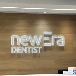K&ouml;nnte beinhalten: Das beleuchtete Schild "new era DENTIST" ist an einer Holzwand angebracht. Das Schild ist wei&szlig; und hat ein modernes Design. Ein wei&szlig;er Empfangstresen ist zu sehen.