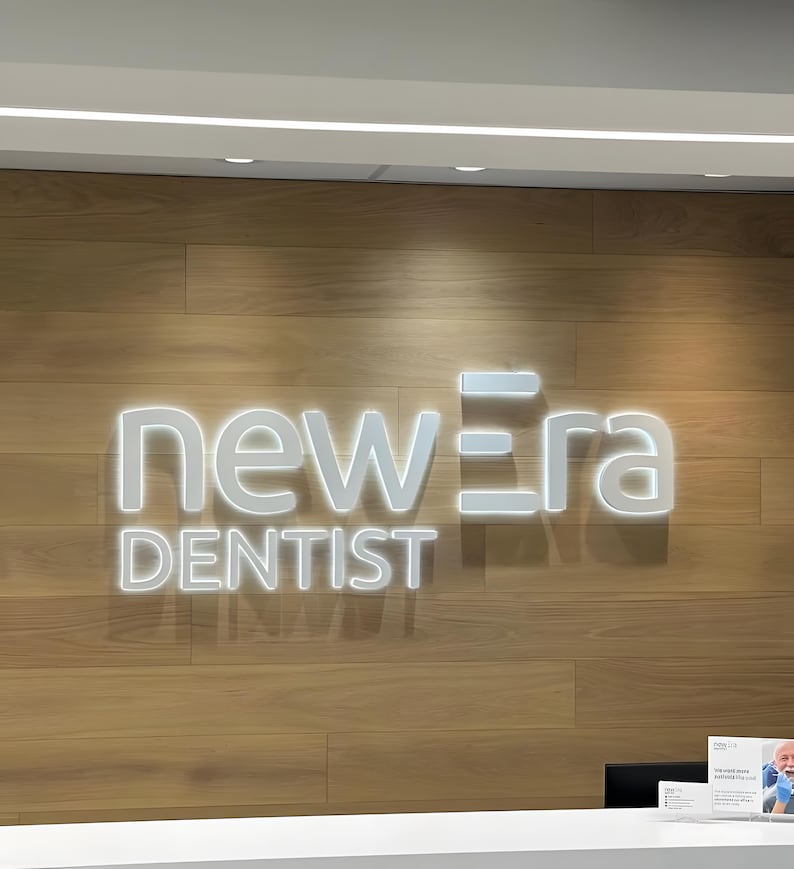 K&ouml;nnte beinhalten: Das beleuchtete Schild "new era DENTIST" ist an einer Holzwand angebracht. Das Schild ist wei&szlig; und hat ein modernes Design. Ein wei&szlig;er Empfangstresen ist zu sehen.