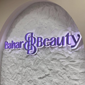 K&ouml;nnte beinhalten: Wandmontiertes Schild mit der Aufschrift "Bahar Beauty" in einer stilisierten Schriftart. Die Buchstaben sind lila mit wei&szlig;em Rand, und das Schild ist an einer strukturierten, hellgrauen Wand angebracht. Das Schild wird von einem weichen, wei&szlig;en Licht beleuchtet.