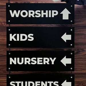 Peut inclure: Panneaux directionnels noirs avec texte et fl&egrave;ches blancs, suspendus par des cha&icirc;nes. Les panneaux indiquent "WORSHIP", "KIDS", "NURSERY" et "STUDENTS". Le fond est un motif de bois fonc&eacute;.