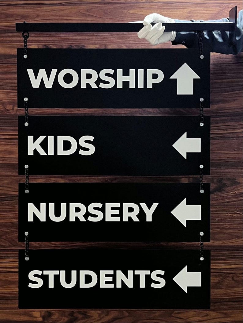 Peut inclure: Panneaux directionnels noirs avec texte et fl&egrave;ches blancs, suspendus par des cha&icirc;nes. Les panneaux indiquent "WORSHIP", "KIDS", "NURSERY" et "STUDENTS". Le fond est un motif de bois fonc&eacute;.