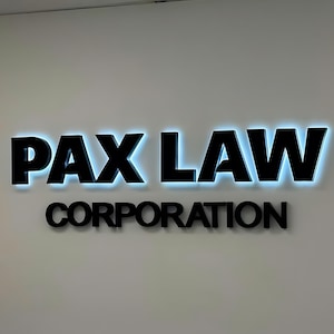 K&ouml;nnte beinhalten: Schwarzes Firmenschild mit der Aufschrift "PAX LAW" und "CORPORATION" an einer wei&szlig;en Wand. Die Buchstaben von "PAX LAW" sind gro&szlig; und fett, mit blauer Hintergrundbeleuchtung. Das Wort "CORPORATION" steht darunter in kleineren Buchstaben.