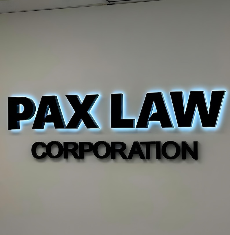 K&ouml;nnte beinhalten: Schwarzes Firmenschild mit der Aufschrift "PAX LAW" und "CORPORATION" an einer wei&szlig;en Wand. Die Buchstaben von "PAX LAW" sind gro&szlig; und fett, mit blauer Hintergrundbeleuchtung. Das Wort "CORPORATION" steht darunter in kleineren Buchstaben.