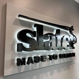 K&ouml;nnte beinhalten: Ein Schild mit dem Namen "Slater" in gro&szlig;en, schwarzen 3D-Buchstaben, mit dem Wort "Slater" in silbernen Buchstaben. Die Worte "MADE TO ORDER" stehen darunter in schwarzen Buchstaben. Das Schild ist auf einem wei&szlig;en Hintergrund montiert.