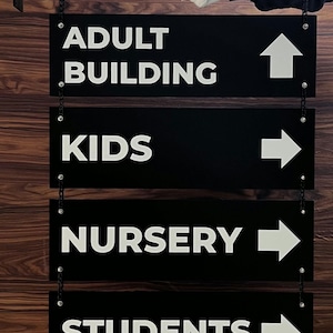 Peut inclure: Panneaux directionnels noirs avec texte et fl&egrave;ches blancs, suspendus par des cha&icirc;nes. Les panneaux indiquent "ADULT BUILDING", "KIDS", "NURSERY" et "STUDENTS". Une main gant&eacute;e de blanc tient le panneau sup&eacute;rieur. Le fond est un grain de bois.