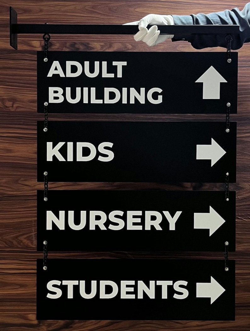 Peut inclure: Panneaux directionnels noirs avec texte et fl&egrave;ches blancs, suspendus par des cha&icirc;nes. Les panneaux indiquent "ADULT BUILDING", "KIDS", "NURSERY" et "STUDENTS". Une main gant&eacute;e de blanc tient le panneau sup&eacute;rieur. Le fond est un grain de bois.