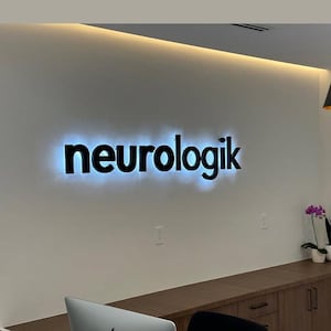 K&ouml;nnte beinhalten: Das Wort "neurologik" wird in schwarzen, beleuchteten Buchstaben vor einer wei&szlig;en Wand angezeigt. Die Buchstaben sind fett und modern, mit einem subtilen blauen Schein, der von hinten ausgeht. Die Szene deutet auf eine professionelle oder medizinische B&uuml;roumgebung hin.