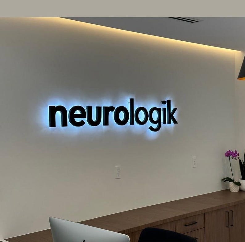 K&ouml;nnte beinhalten: Das Wort "neurologik" wird in schwarzen, beleuchteten Buchstaben vor einer wei&szlig;en Wand angezeigt. Die Buchstaben sind fett und modern, mit einem subtilen blauen Schein, der von hinten ausgeht. Die Szene deutet auf eine professionelle oder medizinische B&uuml;roumgebung hin.