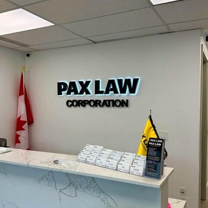 K&ouml;nnte beinhalten: Der Innenraum eines Anwaltsb&uuml;ros zeigt einen wei&szlig;en Empfangstresen mit Marmormuster. An der Wand steht der Text "PAX LAW CORPORATION" in schwarzen Buchstaben mit blauer Hintergrundbeleuchtung. Links h&auml;ngt eine kanadische Flagge.