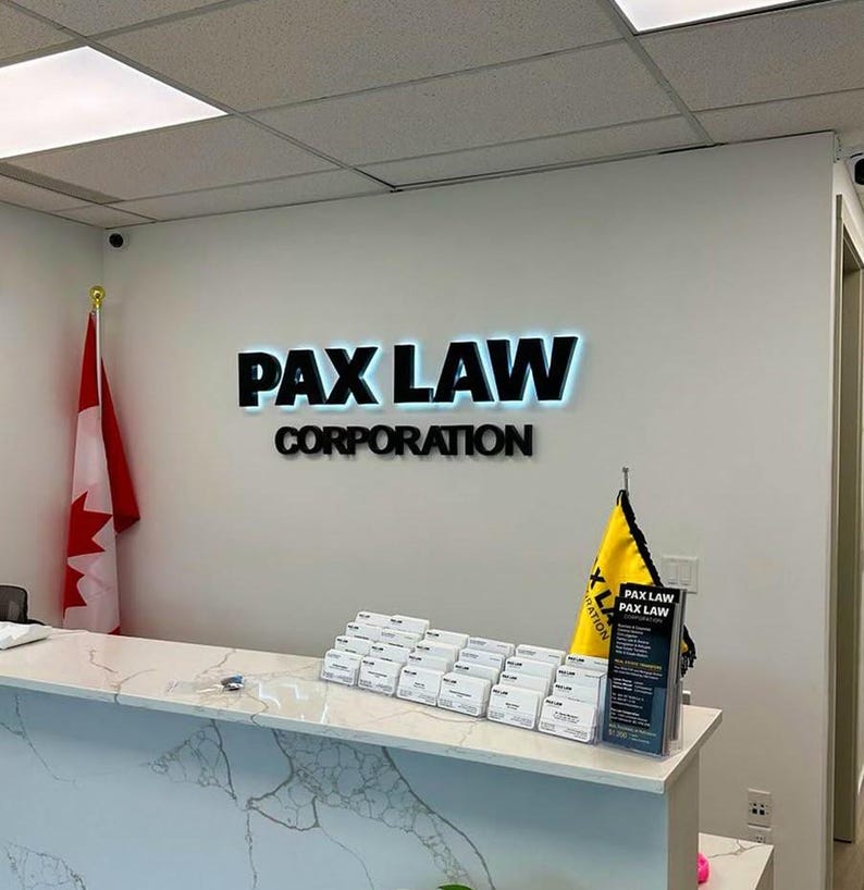 K&ouml;nnte beinhalten: Der Innenraum eines Anwaltsb&uuml;ros zeigt einen wei&szlig;en Empfangstresen mit Marmormuster. An der Wand steht der Text "PAX LAW CORPORATION" in schwarzen Buchstaben mit blauer Hintergrundbeleuchtung. Links h&auml;ngt eine kanadische Flagge.